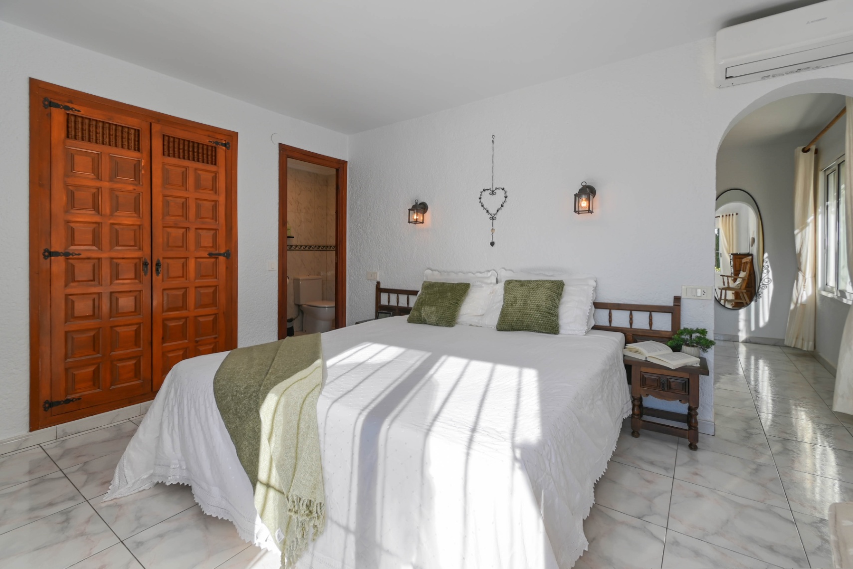 Bedroom featuring a carved wooden built-in closet, light marble floors, and an ensuite bathroom. Natural light illuminates the bed styled with green accents and lantern wall sconces.