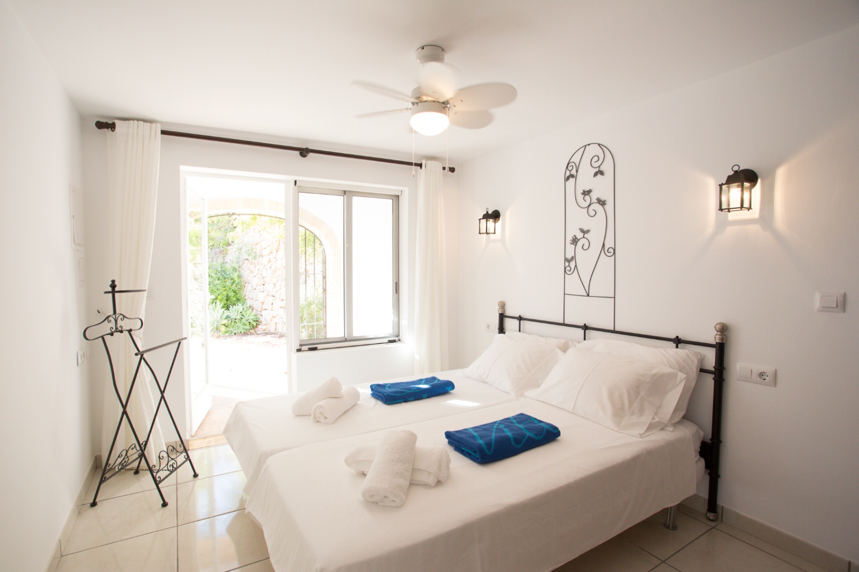 Bedroom featuring twin beds, a wrought iron headboard, and wall lanterns. Includes a ceiling fan and direct access to a stone-walled patio through a glass door.