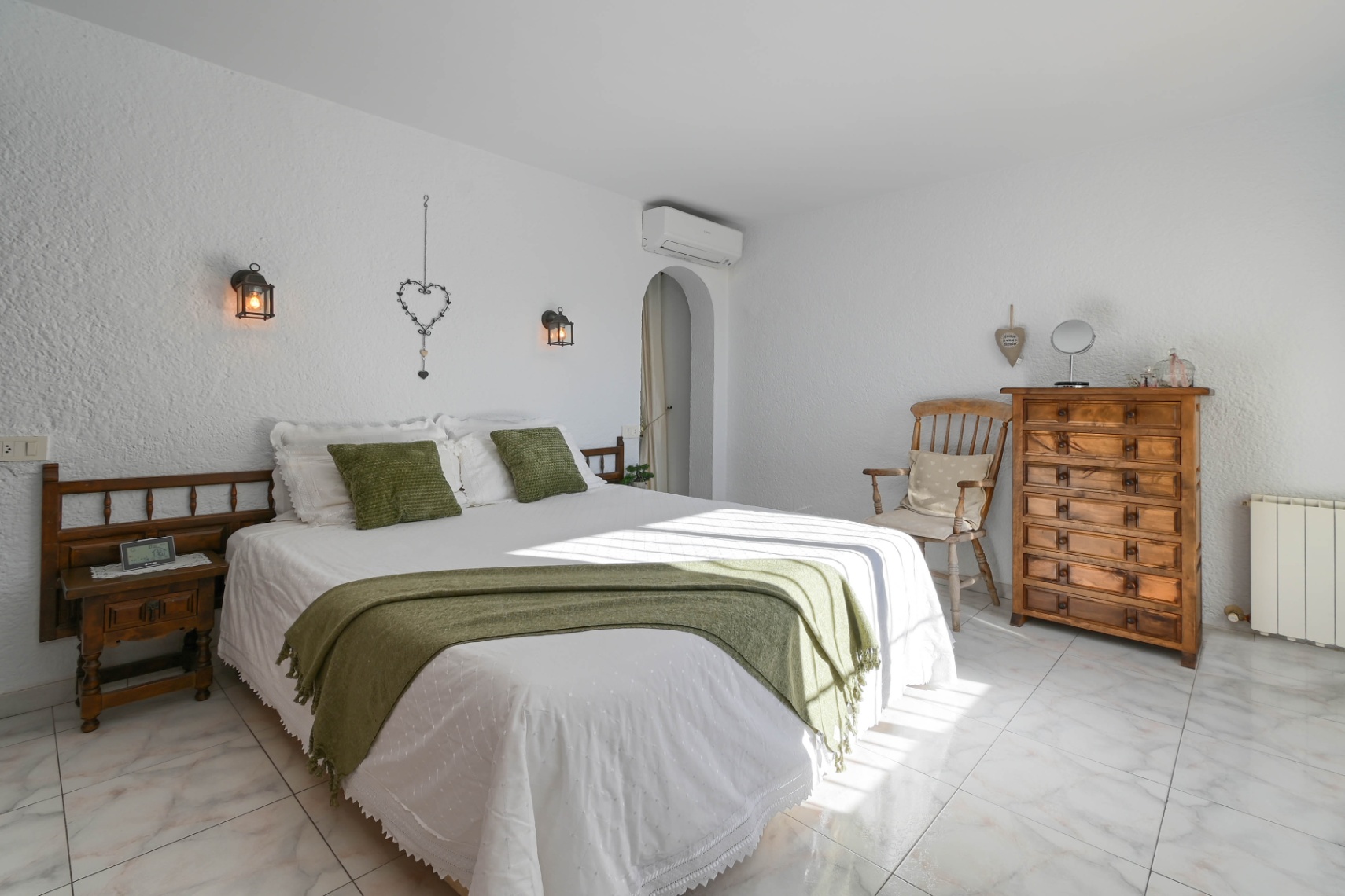 Bedroom with white textured walls, marble flooring, and rustic wooden furniture. Features natural light, green accents, and wall-mounted lantern lighting.