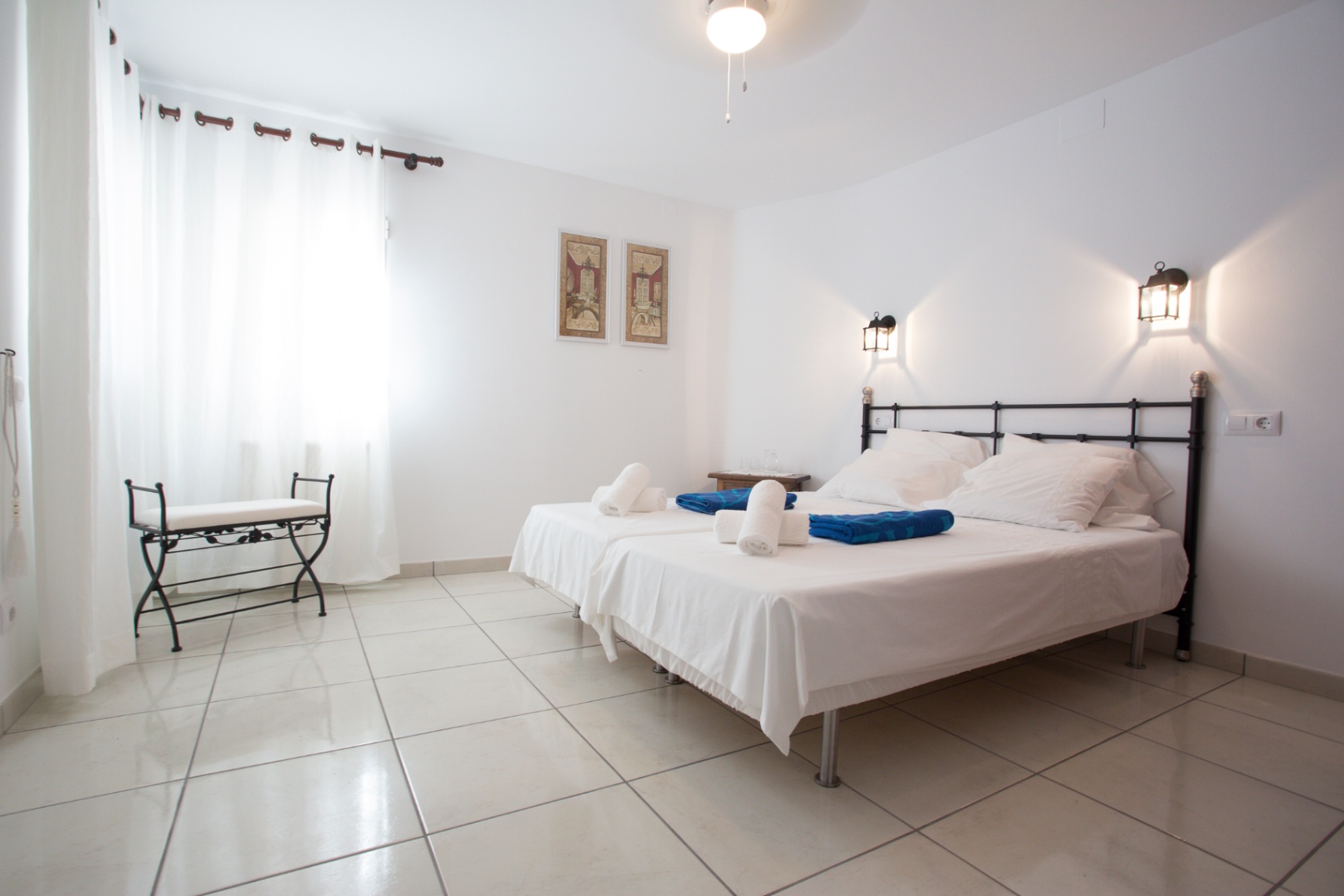 Bright bedroom featuring twin beds with a wrought iron headboard, tiled flooring, and wall-mounted lantern lighting. A matching iron bench sits by the window.