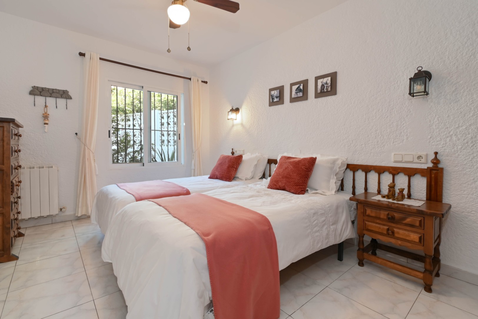 Bright bedroom featuring twin beds with terracotta accents, rustic wooden furniture, and textured white walls. A window with security bars offers views of the garden.