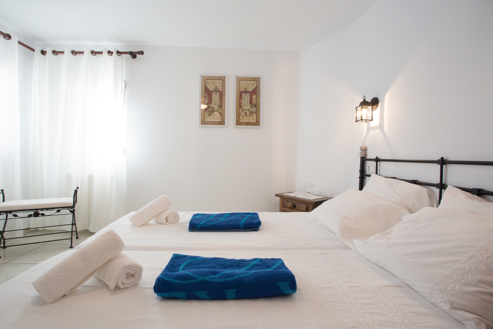 Bright bedroom featuring twin beds with white linens, a black wrought iron headboard, and a matching bench. Natural light filters through white curtains.