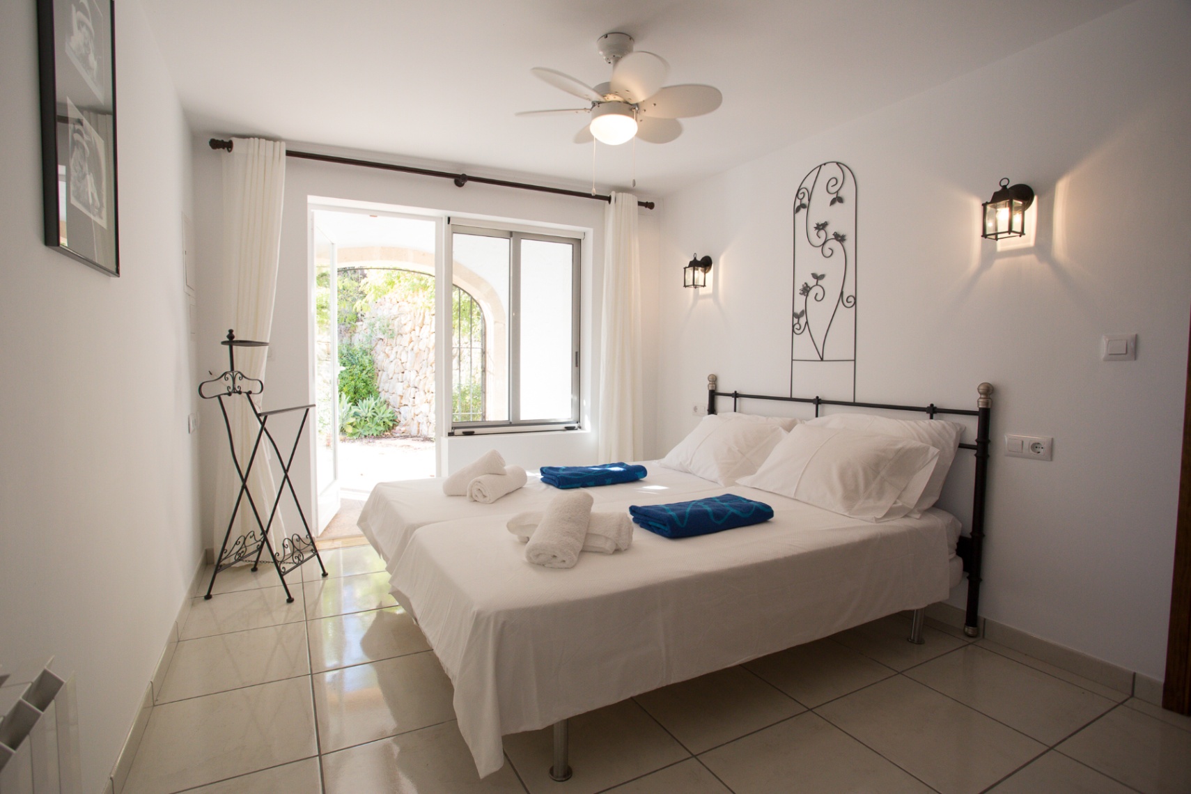 Bright bedroom featuring twin beds, wrought iron accents, and a ceiling fan. Large glass doors provide direct access to a private stone-walled patio.