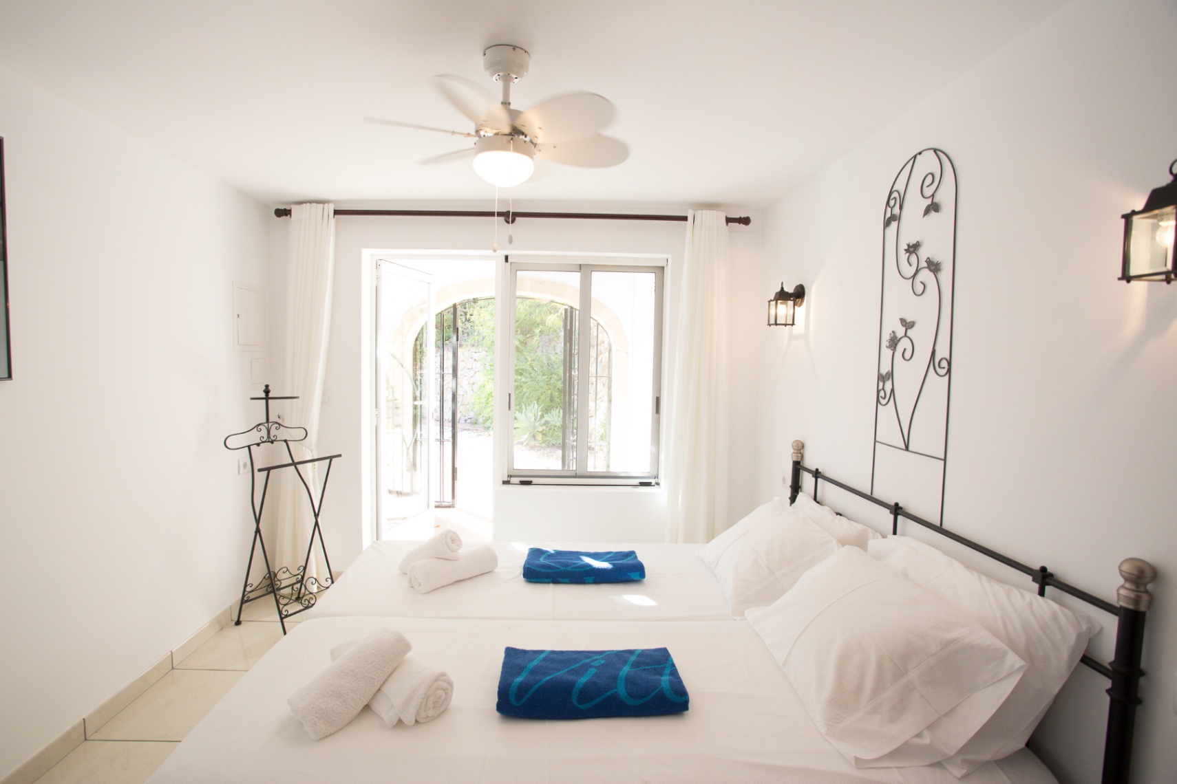 Bright bedroom featuring white walls, a wrought iron bed frame, and a ceiling fan. Glass doors open directly onto a sunlit patio with garden views.