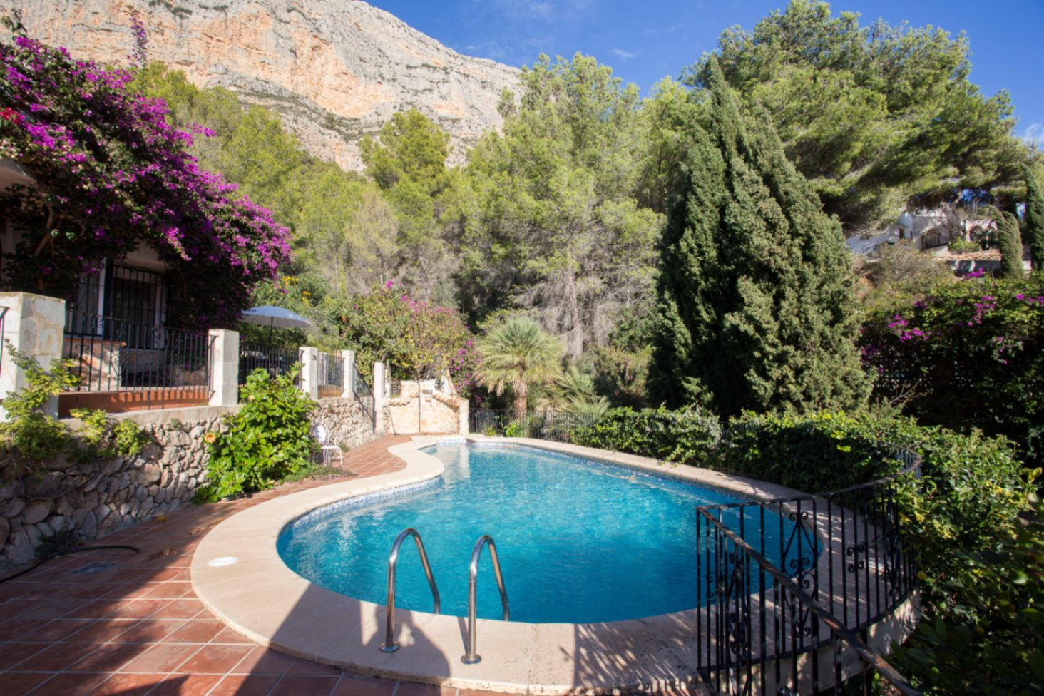 Curved outdoor pool surrounded by terracotta tiling and lush Mediterranean vegetation, set against a dramatic limestone cliff backdrop.