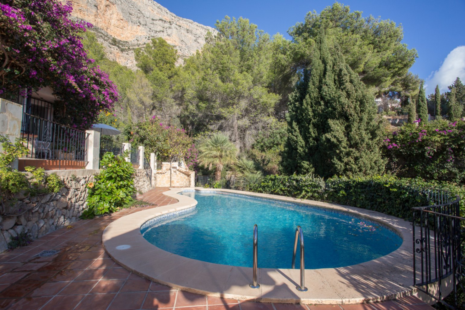 Curved swimming pool with blue mosaic tiling, surrounded by terracotta paving and lush Mediterranean greenery against a dramatic mountain backdrop.