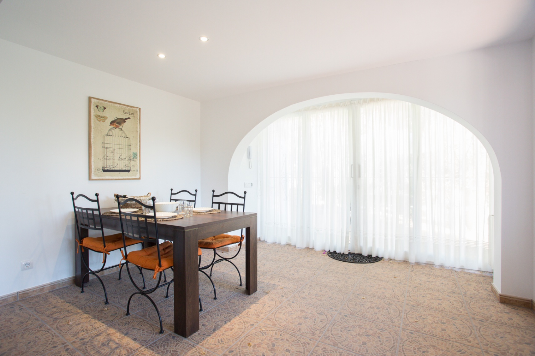 Dining area featuring a dark wood table and wrought iron chairs. A large arched opening with sheer curtains provides natural light over the patterned tile flooring.