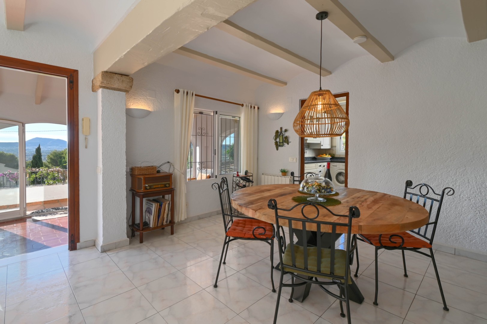 Dining area featuring a round wooden table, wrought iron chairs, and a rattan pendant lamp. Includes exposed ceiling beams and views toward a terrace and distant mountains.