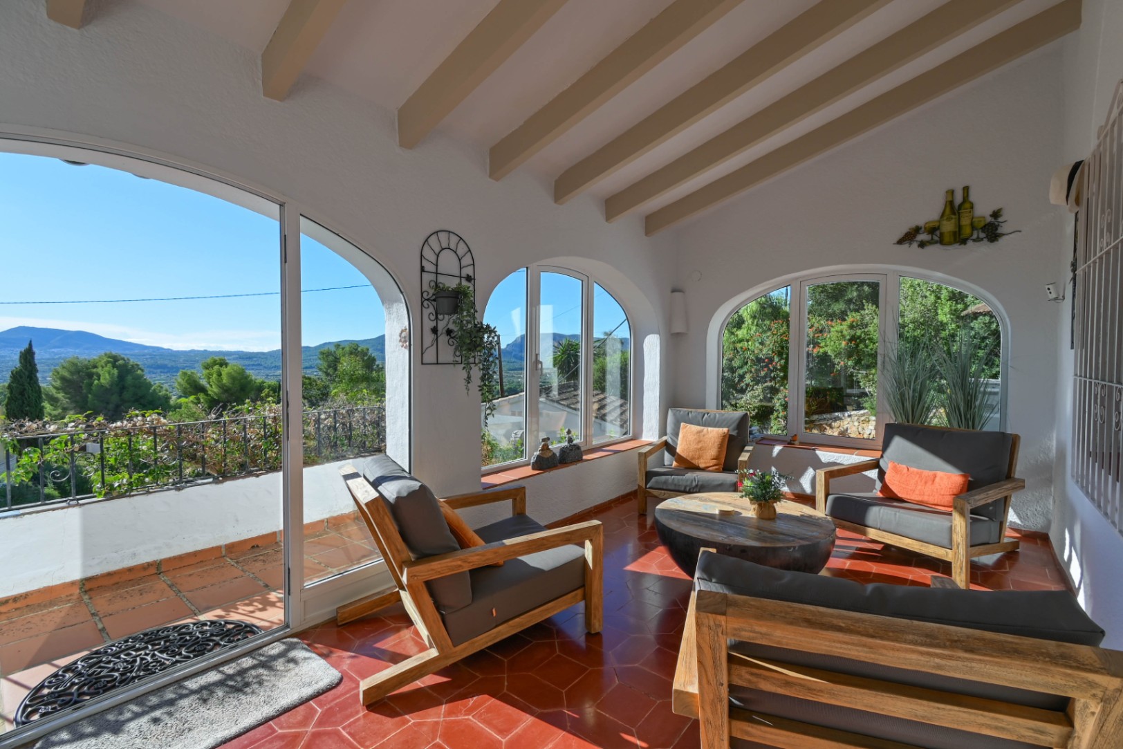 Enclosed terrace featuring exposed wooden beams, hexagonal terracotta flooring, and arched windows with panoramic views of the mountains.