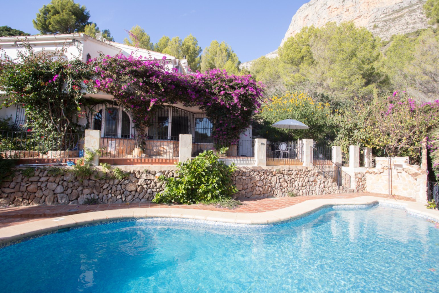 Exterior view featuring a curved swimming pool, terracotta terrace, and a white villa draped in purple bougainvillea against a limestone mountain backdrop.