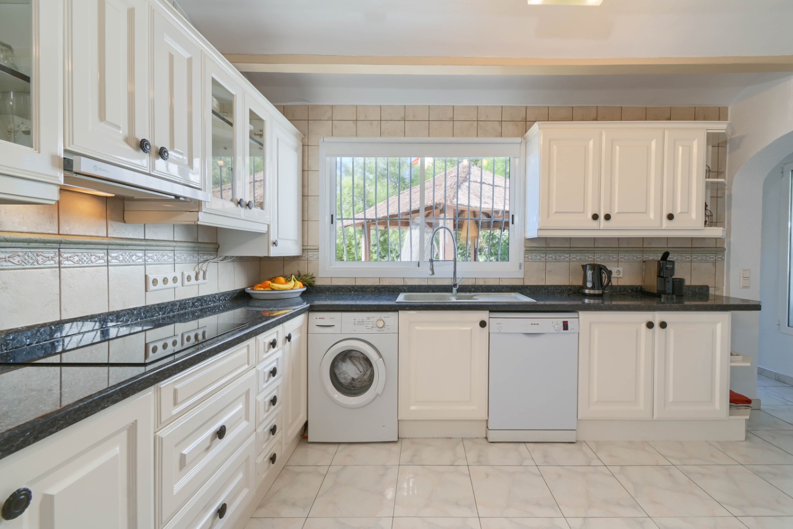 Functional kitchen featuring white cabinetry, dark granite countertops, and integrated appliances. Includes a laundry area and a window with garden views.