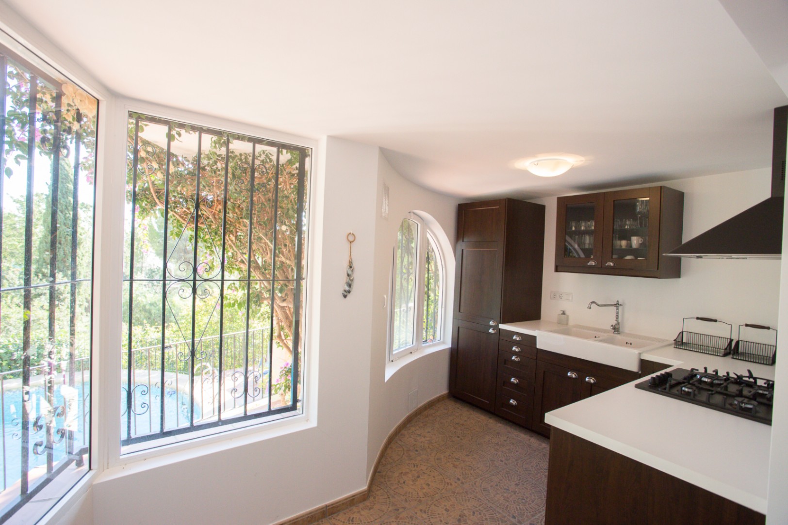 Kitchen featuring dark wood cabinetry, white countertops, and a farmhouse sink. Large windows with decorative ironwork overlook the garden and swimming pool.