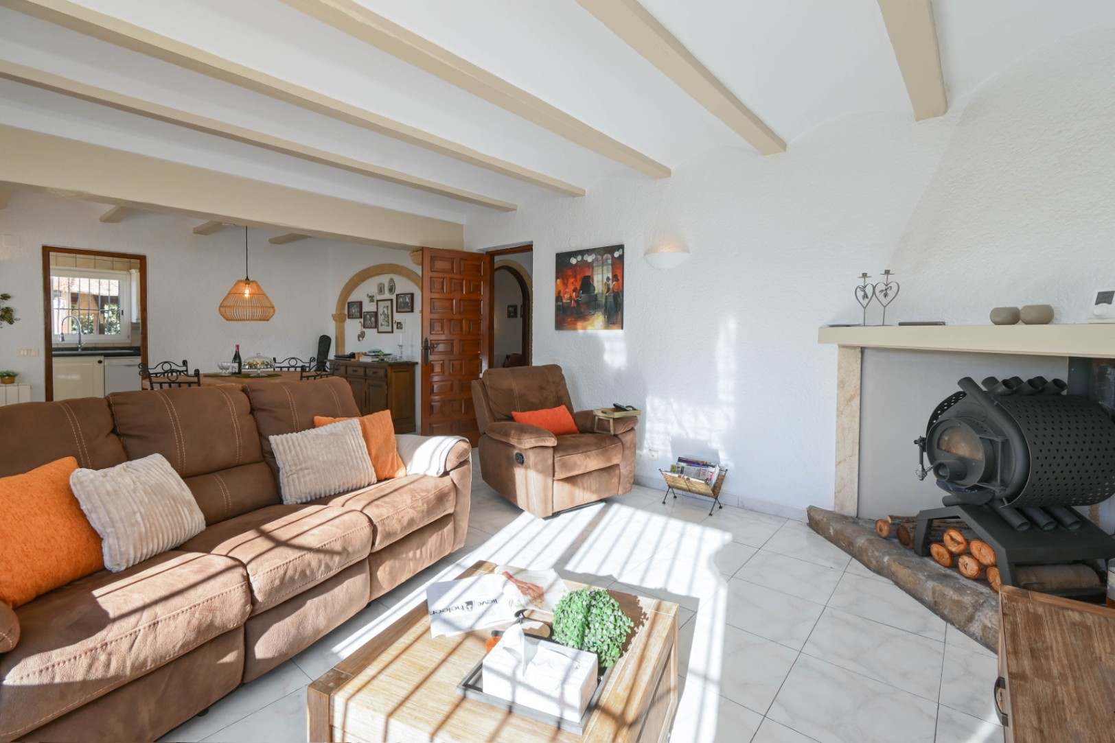 Living room with exposed ceiling beams, a brown fabric sofa, and a traditional wood-burning stove set in a fireplace alcove. Natural light highlights the tiled flooring.
