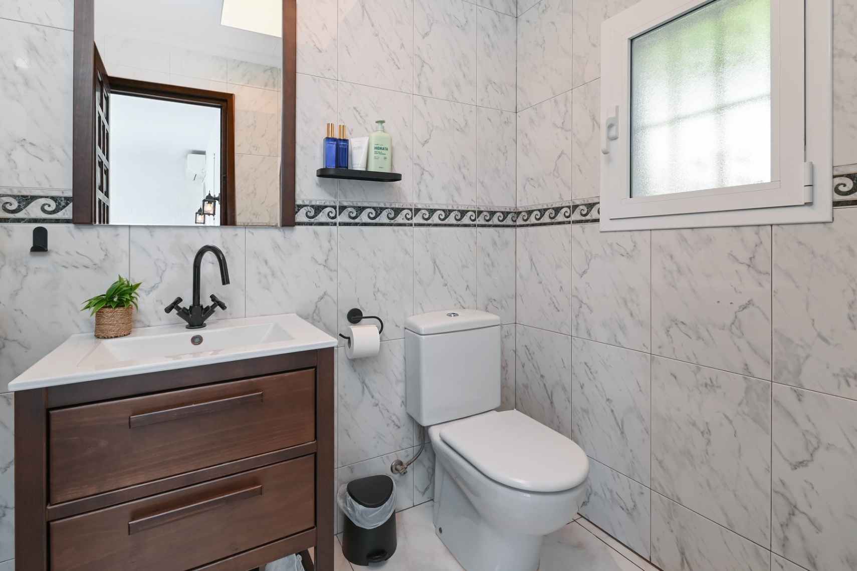 Modern bathroom featuring marble-patterned wall tiles, a dark wood vanity with a white sink, matte black fixtures, and a frosted glass window for privacy.