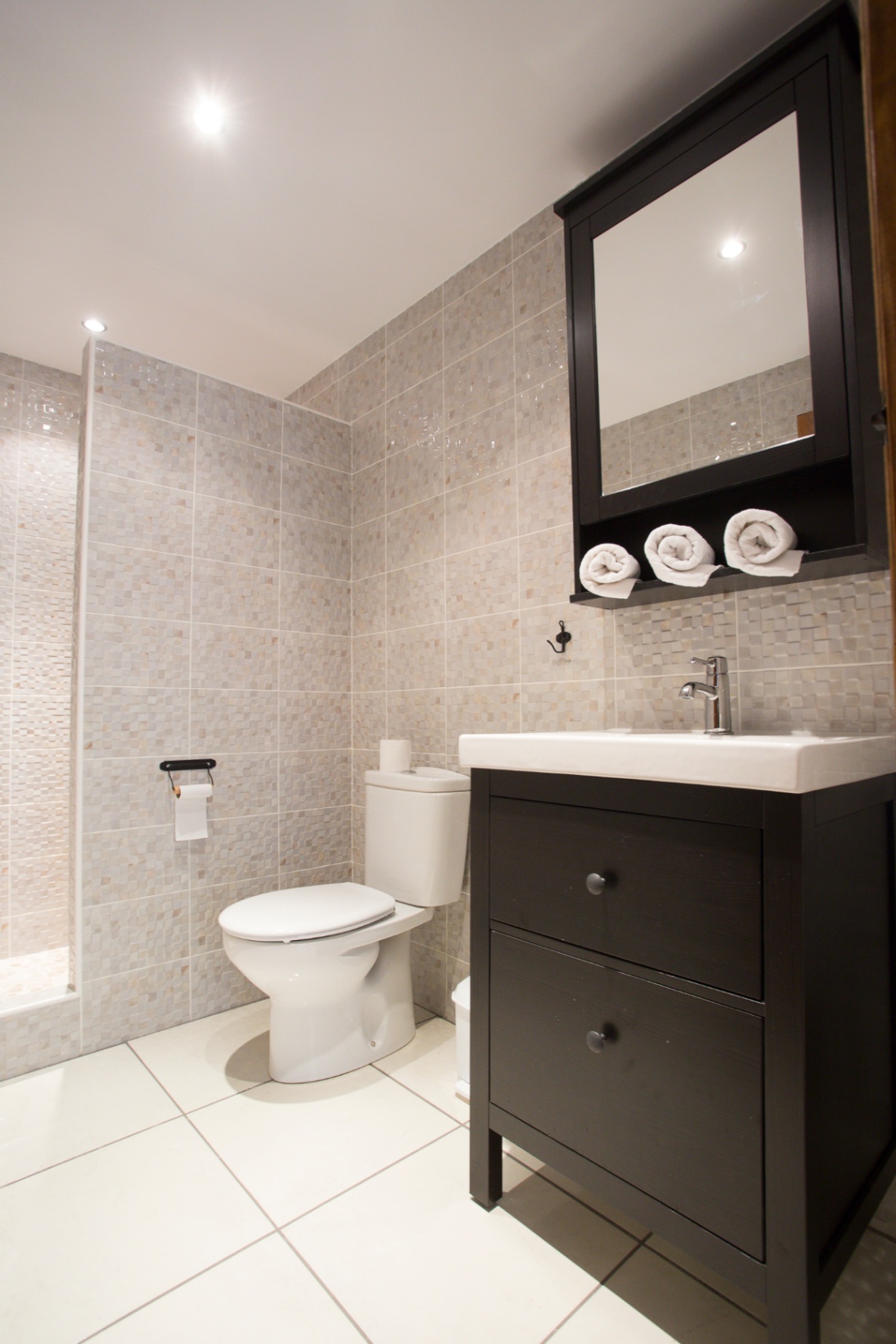 Modern bathroom featuring neutral mosaic wall tiles, a dark wood vanity with a white ceramic sink, and matching mirrored cabinet with integrated towel storage.
