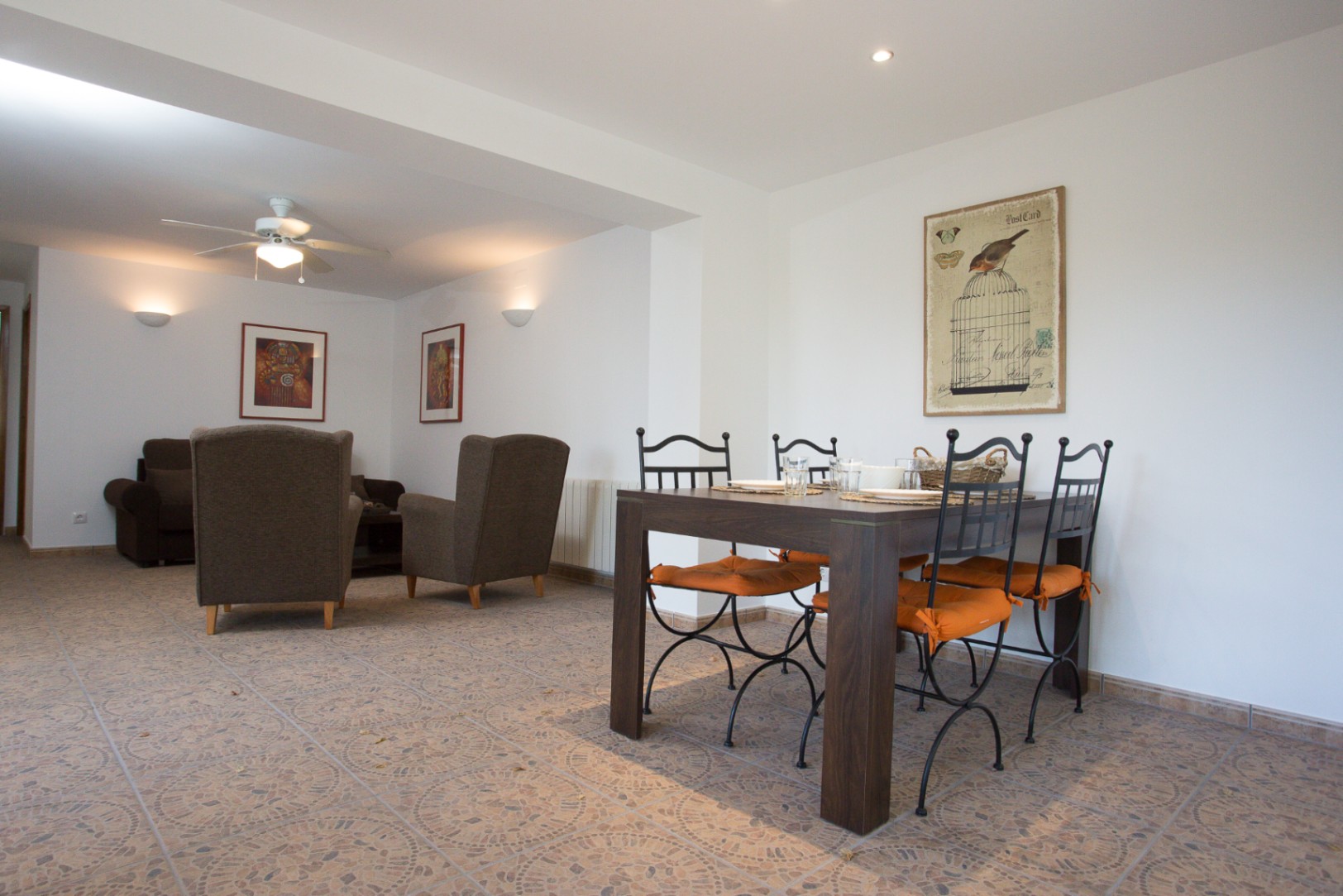 Open-plan living space featuring a dark wood dining table with wrought-iron chairs and orange cushions, set against patterned tile flooring and a neutral seating area.
