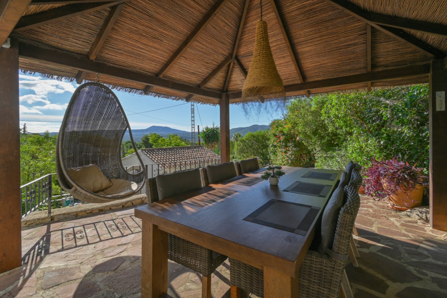Outdoor dining area under a thatched gazebo featuring a wooden table, wicker seating, and a hanging egg chair with views of the surrounding mountains.