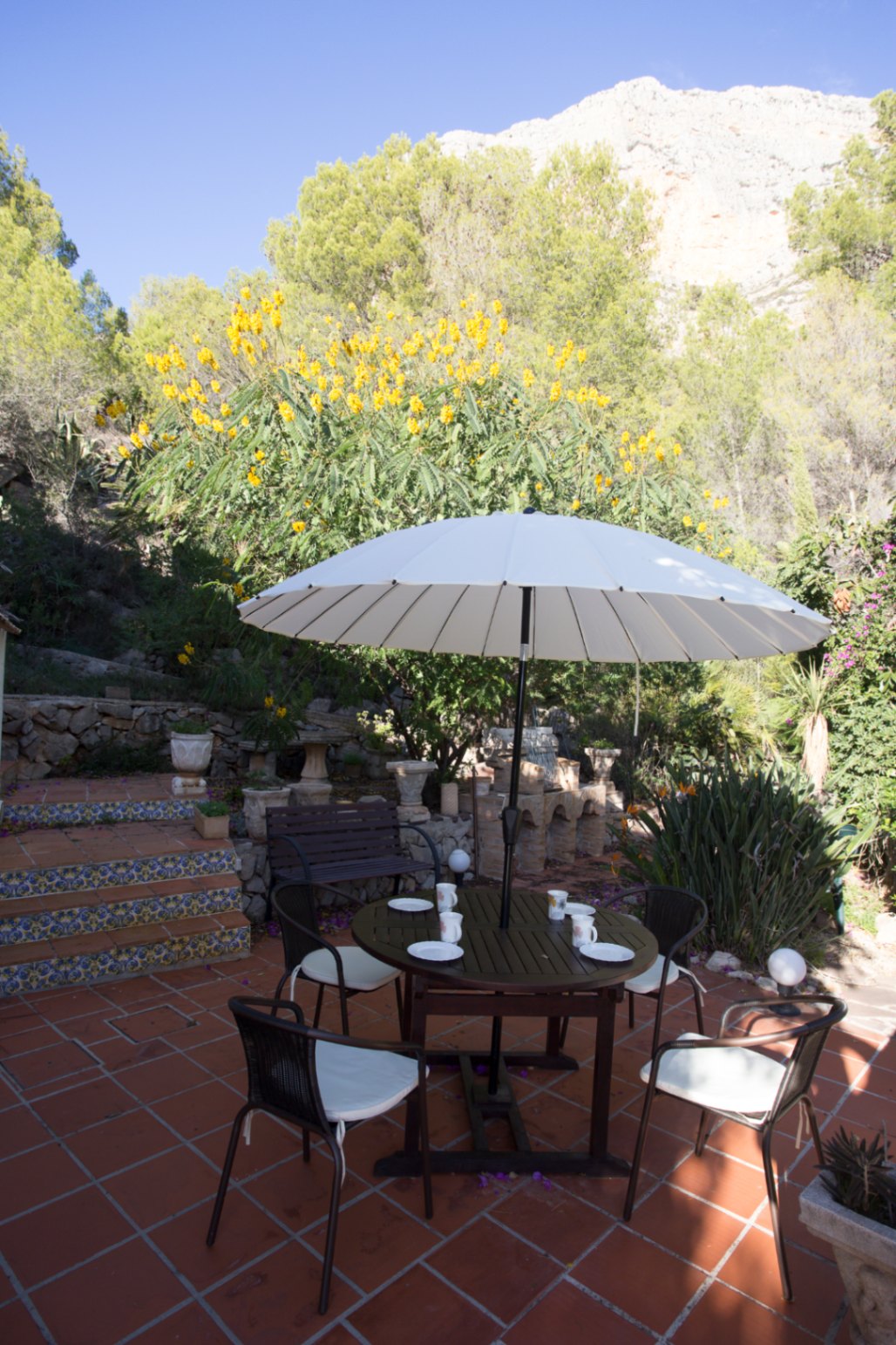 Outdoor terrace with terracotta flooring, a round dining table under a white parasol, and decorative tiled steps leading to a lush garden with mountain views.