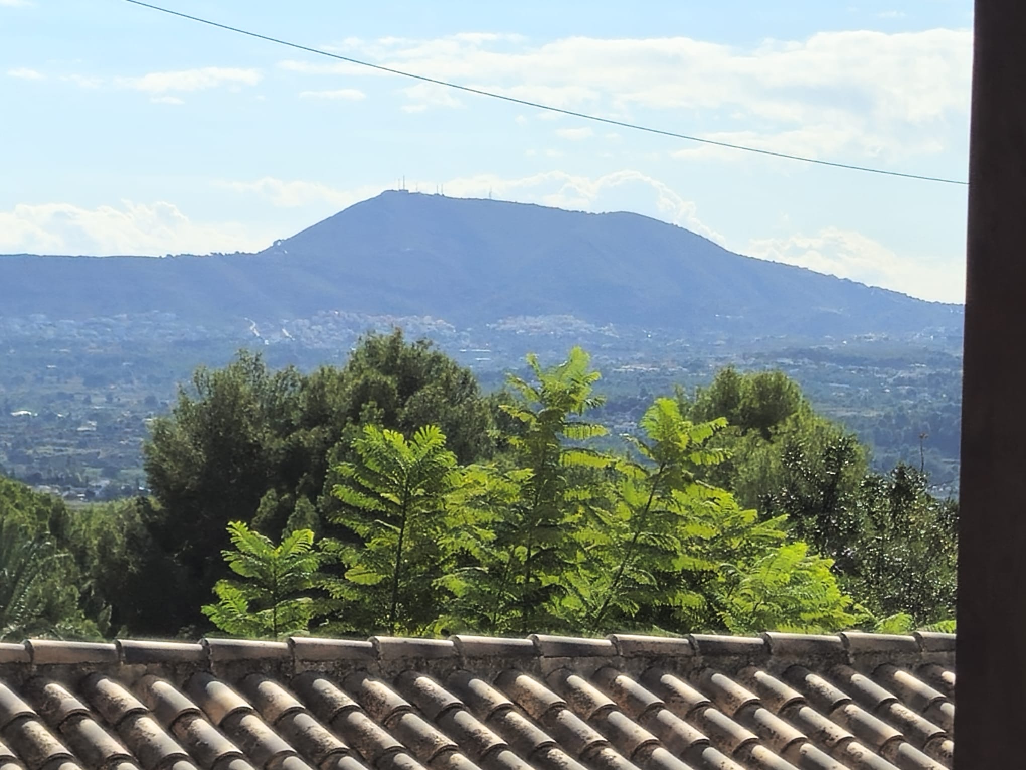Panoramic view from the rooftop featuring traditional clay tiles, lush green foliage, and a prominent mountain peak on the horizon under a clear sky.