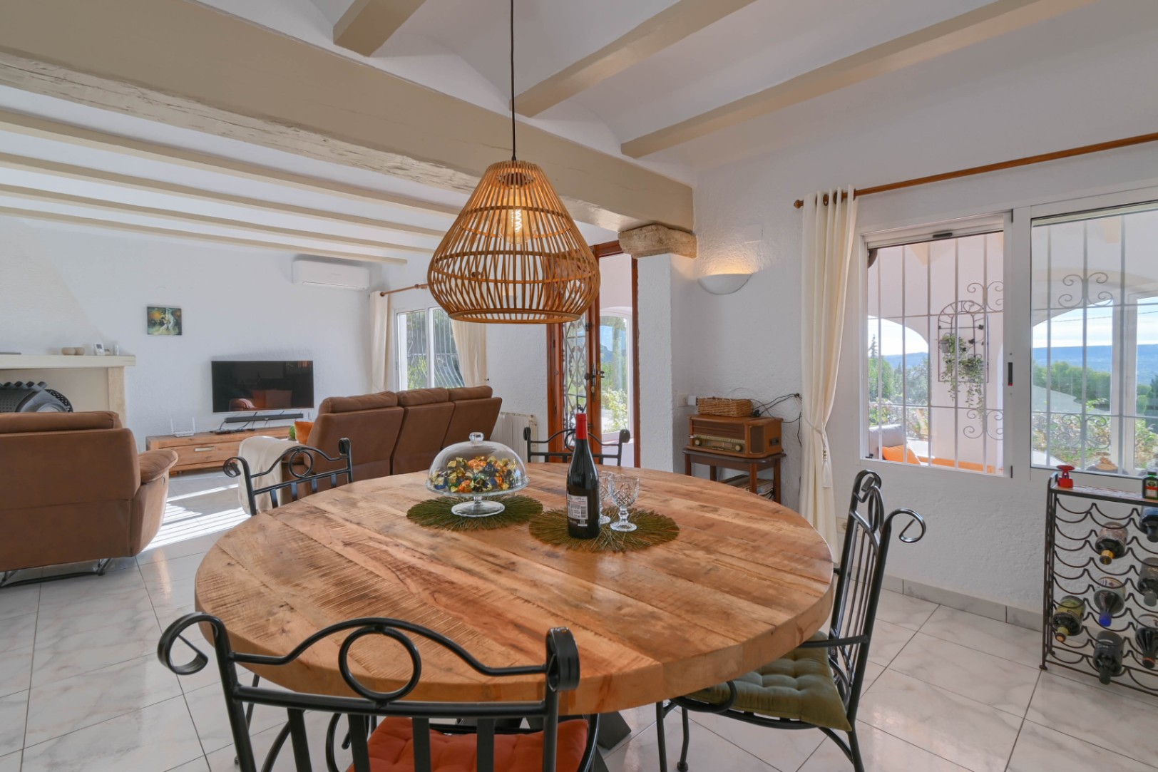 Round wooden dining table with wrought iron chairs under a wicker pendant lamp, featuring exposed ceiling beams and a view into the living area.