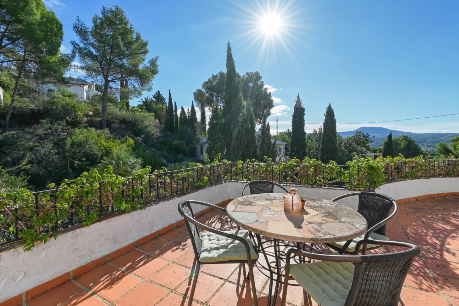 Terrace featuring terracotta tiling, a stone-top table, and wicker seating, overlooking Mediterranean greenery and mountains under direct sunlight.