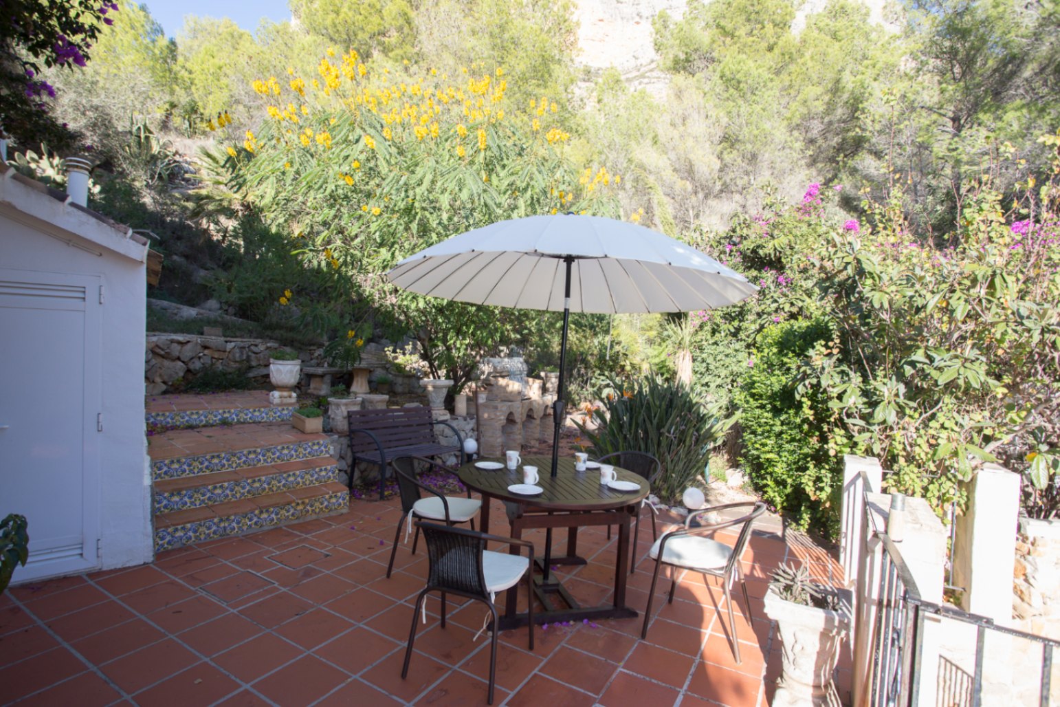 Terrace with terracotta flooring, featuring a wooden dining set under a white umbrella, bordered by patterned tile steps and lush Mediterranean hillside vegetation.