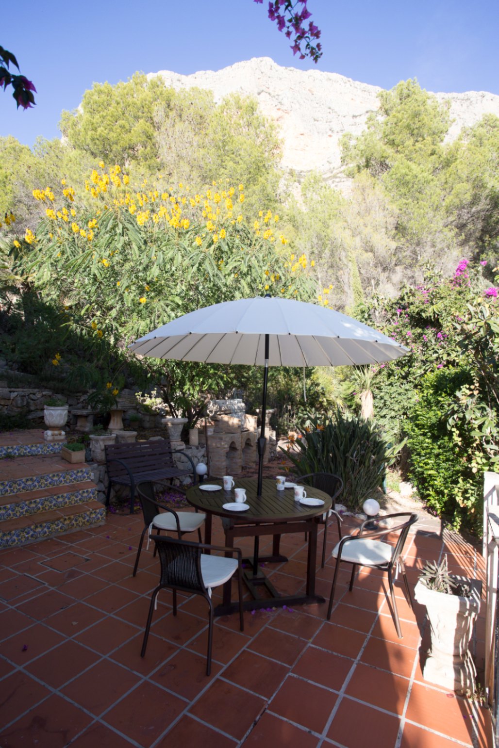 Terrace with terracotta tiles featuring a circular wooden dining set and white parasol, set against a backdrop of Mediterranean flora and rocky cliffs.