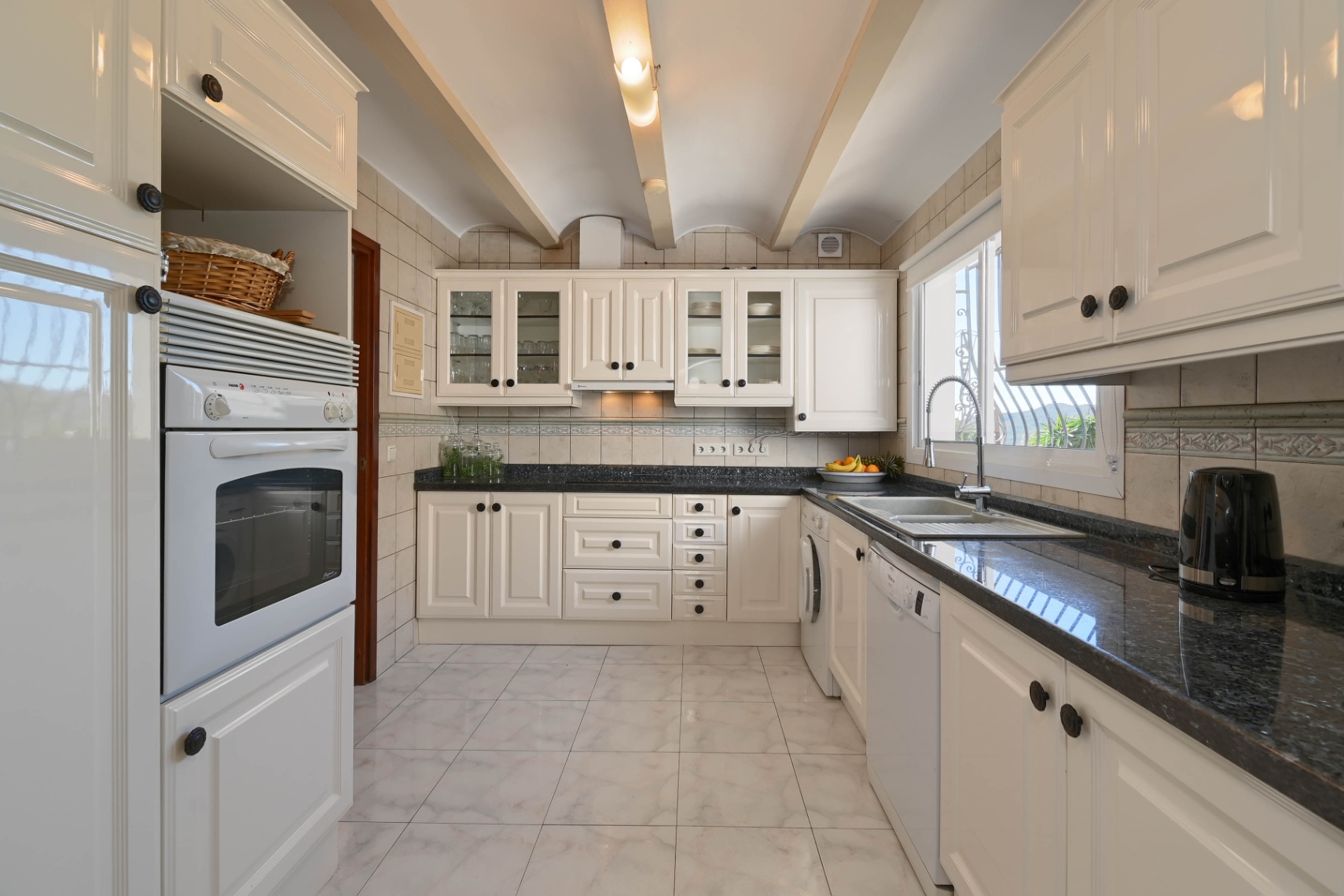 Traditional kitchen featuring cream cabinetry, dark granite countertops, and a vaulted ceiling with exposed beams and integrated appliances.