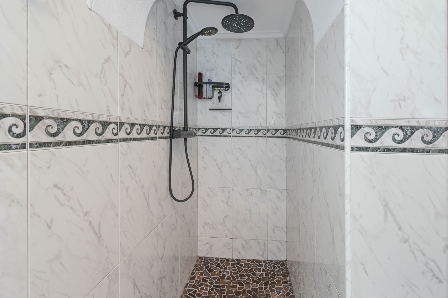 Walk-in shower featuring marble-effect wall tiles with a decorative wave border, a pebble stone floor, and a modern matte black rain shower system.