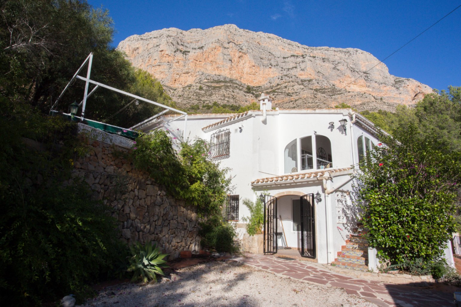 White Mediterranean villa with arched windows and stone accents, situated at the foot of a prominent rocky mountain under a clear sky.