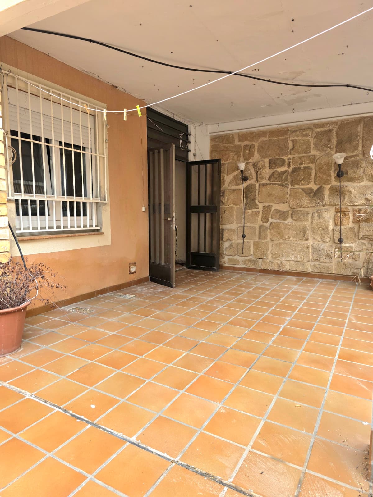 Covered patio featuring terracotta tile flooring, a rustic stone accent wall with wall-mounted sconces, and a window with white security bars.