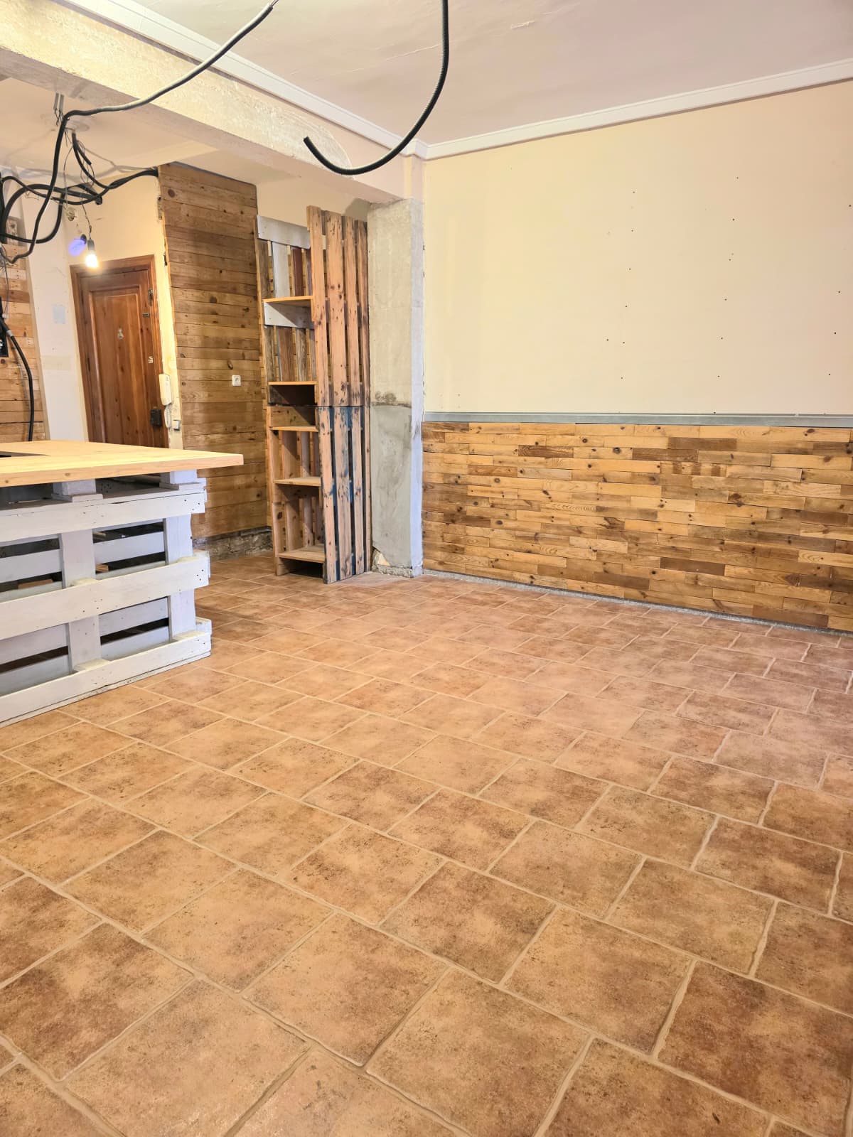 Interior space featuring terracotta tile flooring and rustic wood-slat wall paneling. A white pallet-based counter and wooden shelving define the functional layout.