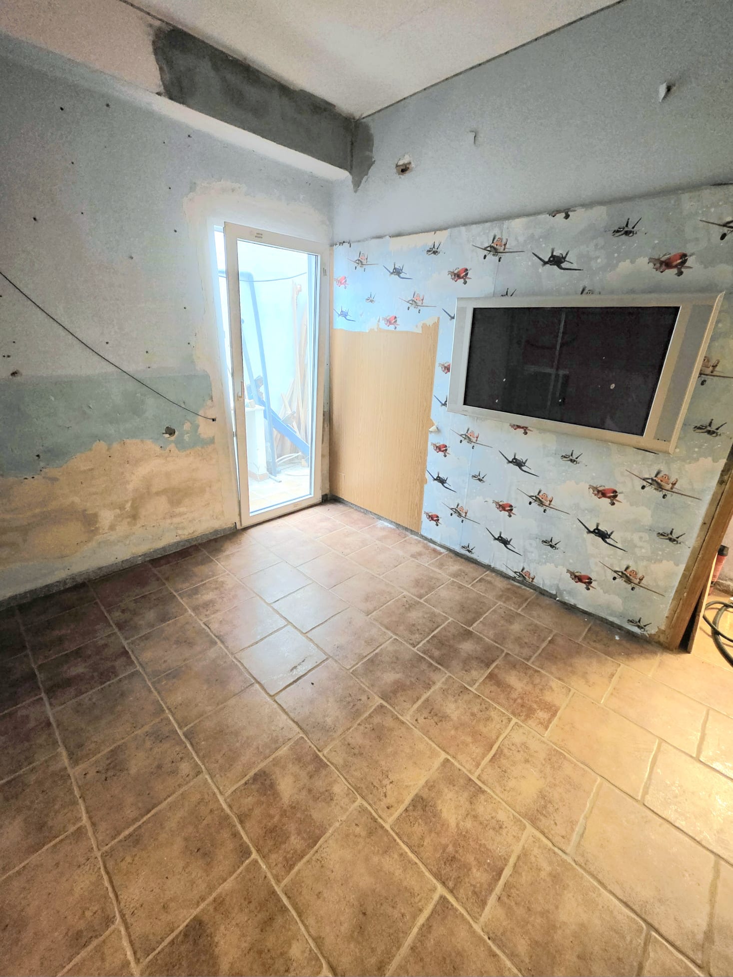 Interior space under renovation with textured ceramic floor tiles, a white-framed glass door, and a feature wall with airplane-patterned wallpaper and a built-in monitor.