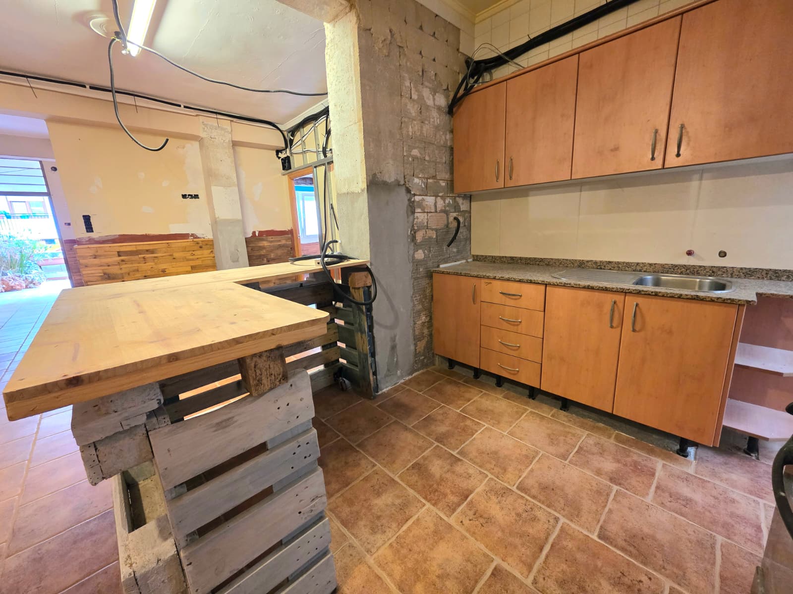 Kitchen space undergoing renovation with wooden cabinets, granite countertops, terracotta tiling, and a makeshift pallet island under exposed wiring.