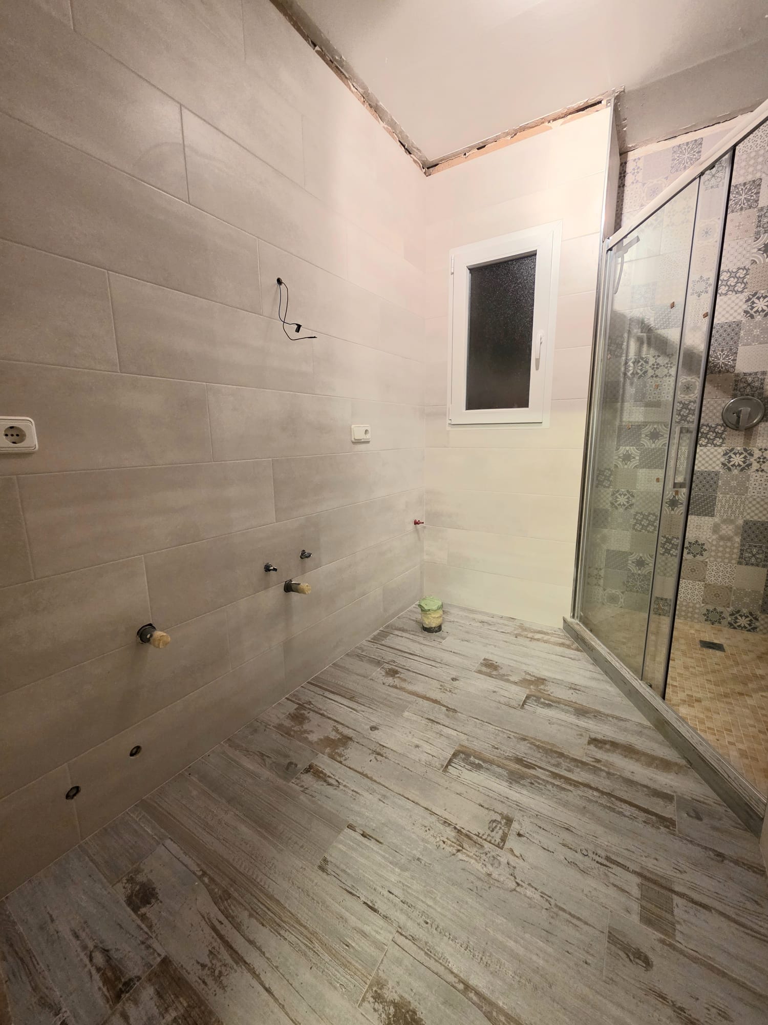Modern bathroom under renovation featuring wood-effect floor tiles, large format wall tiling, and a glass shower enclosure with decorative patterned tiles.