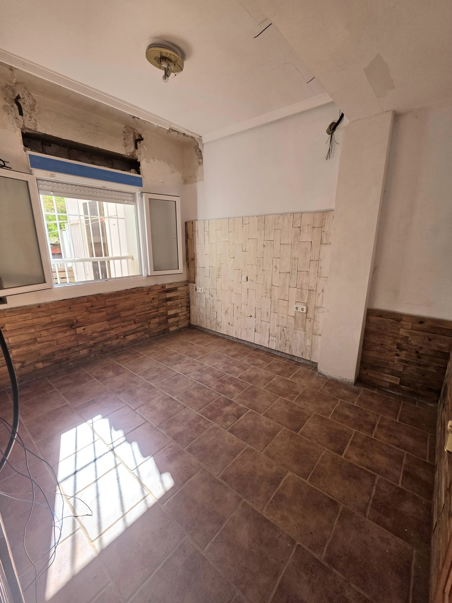 Room featuring terracotta tile flooring and decorative wood-effect wall cladding. Natural light enters through a white-framed window, highlighting renovation details.