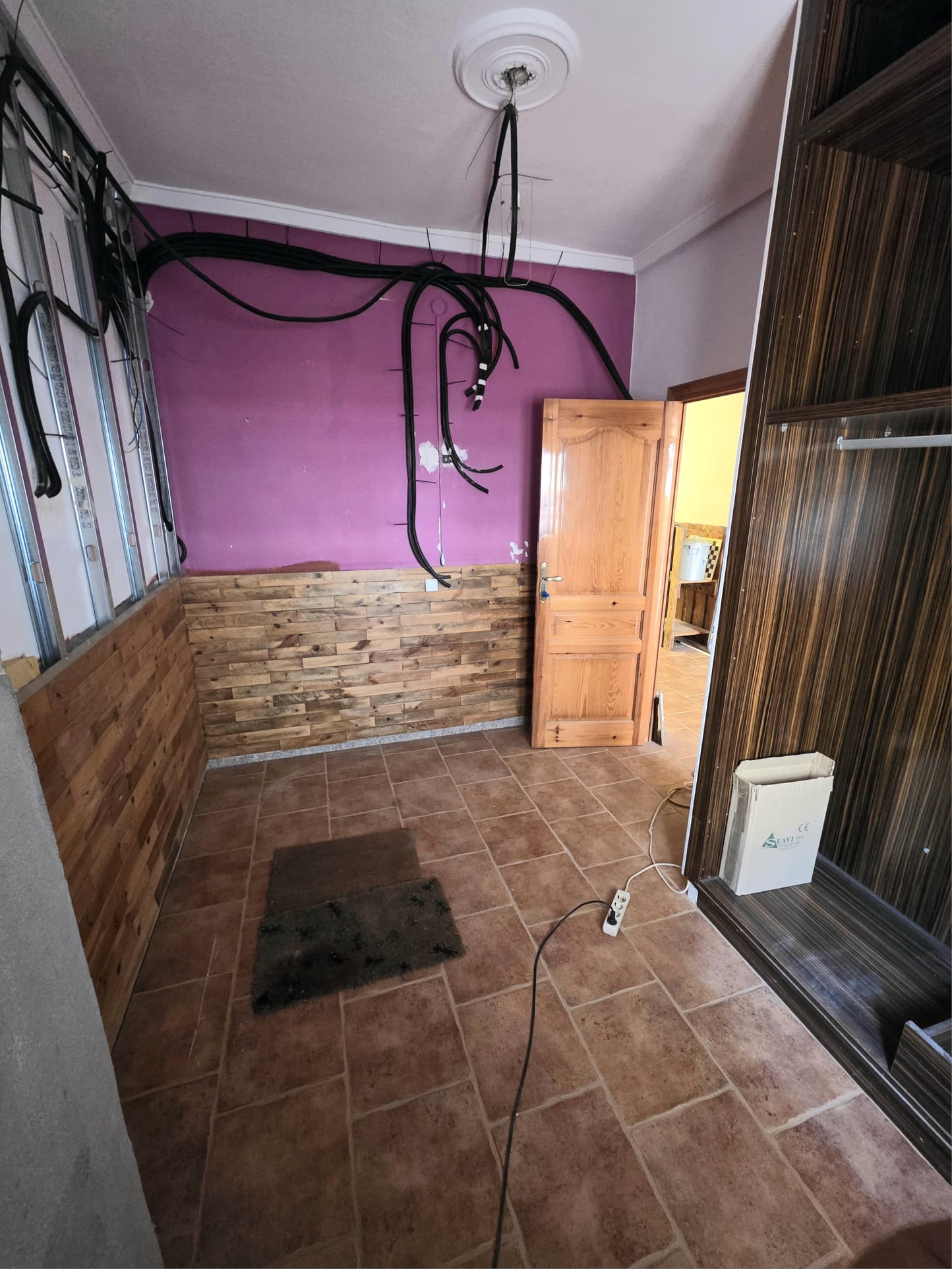 Room under renovation featuring terracotta tile flooring, purple walls with wooden cladding, and exposed electrical wiring and metal framing.