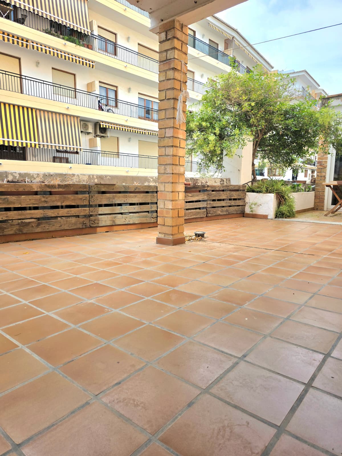 Spacious outdoor terrace featuring terracotta tile flooring, a central brick pillar, and custom wooden pallet fencing alongside integrated greenery.