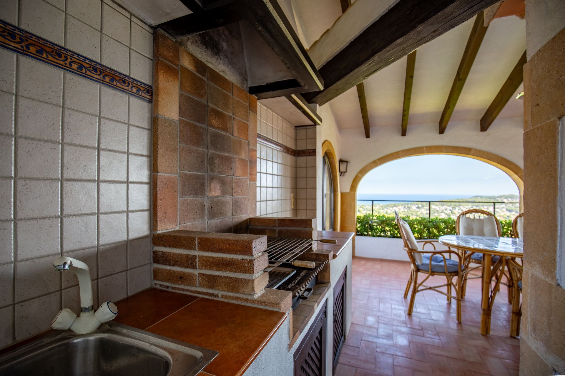 Covered outdoor kitchen featuring a built-in brick barbecue, sink, and terracotta flooring, opening onto a terrace with sea views and exposed wooden beams.