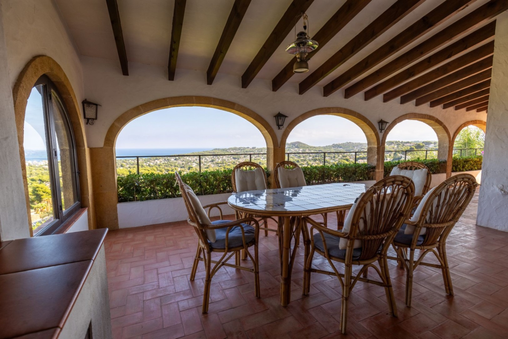 Covered terrace featuring stone arches, exposed wooden beams, and terracotta flooring. Includes a wicker dining set with panoramic views of the coastline and hills.