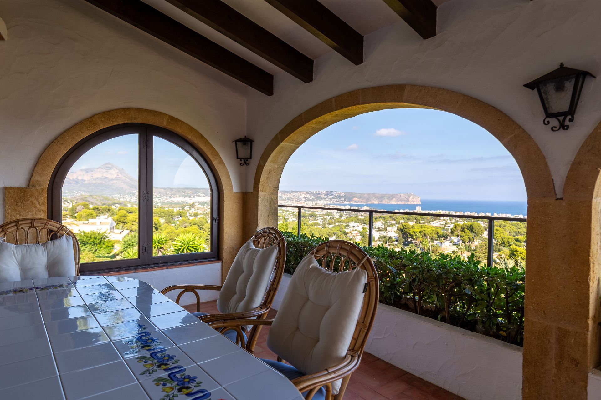 Covered terrace featuring stone arches, wooden beams, and a tiled dining table. Panoramic views of the Mediterranean coast and mountains are visible from the seating area.