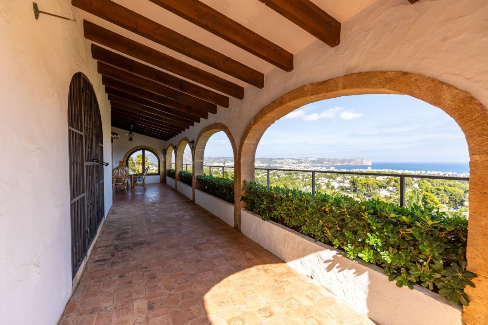 Covered terrace featuring traditional stone arches, exposed dark wood ceiling beams, and terracotta flooring. Integrated planters frame panoramic coastal and sea views.