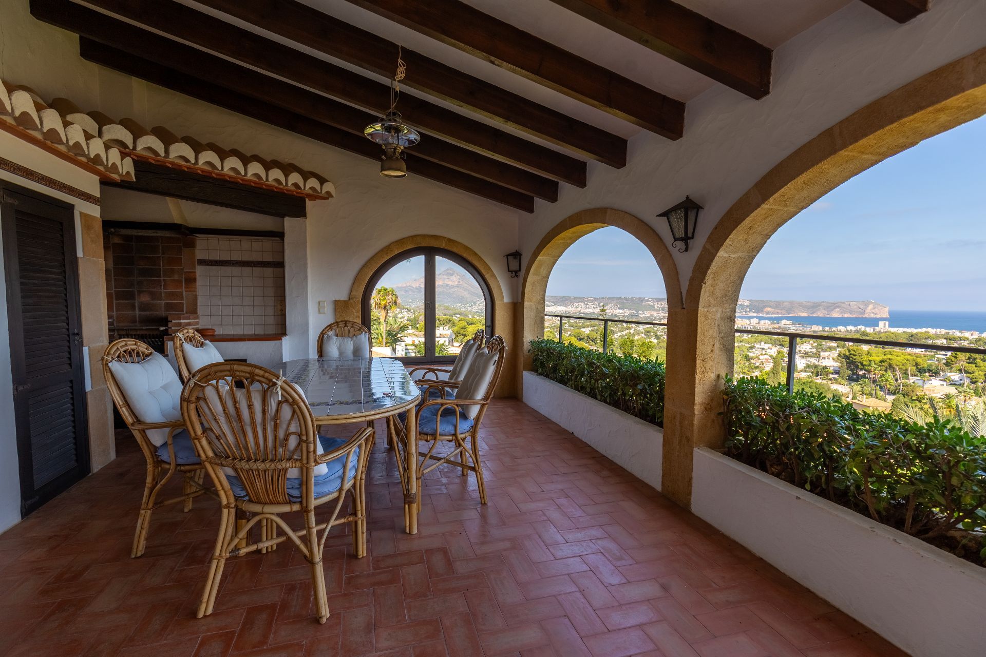 Covered terrace with stone arches, wooden beams, and terracotta flooring. Features a dining area with panoramic coastal and mountain views.