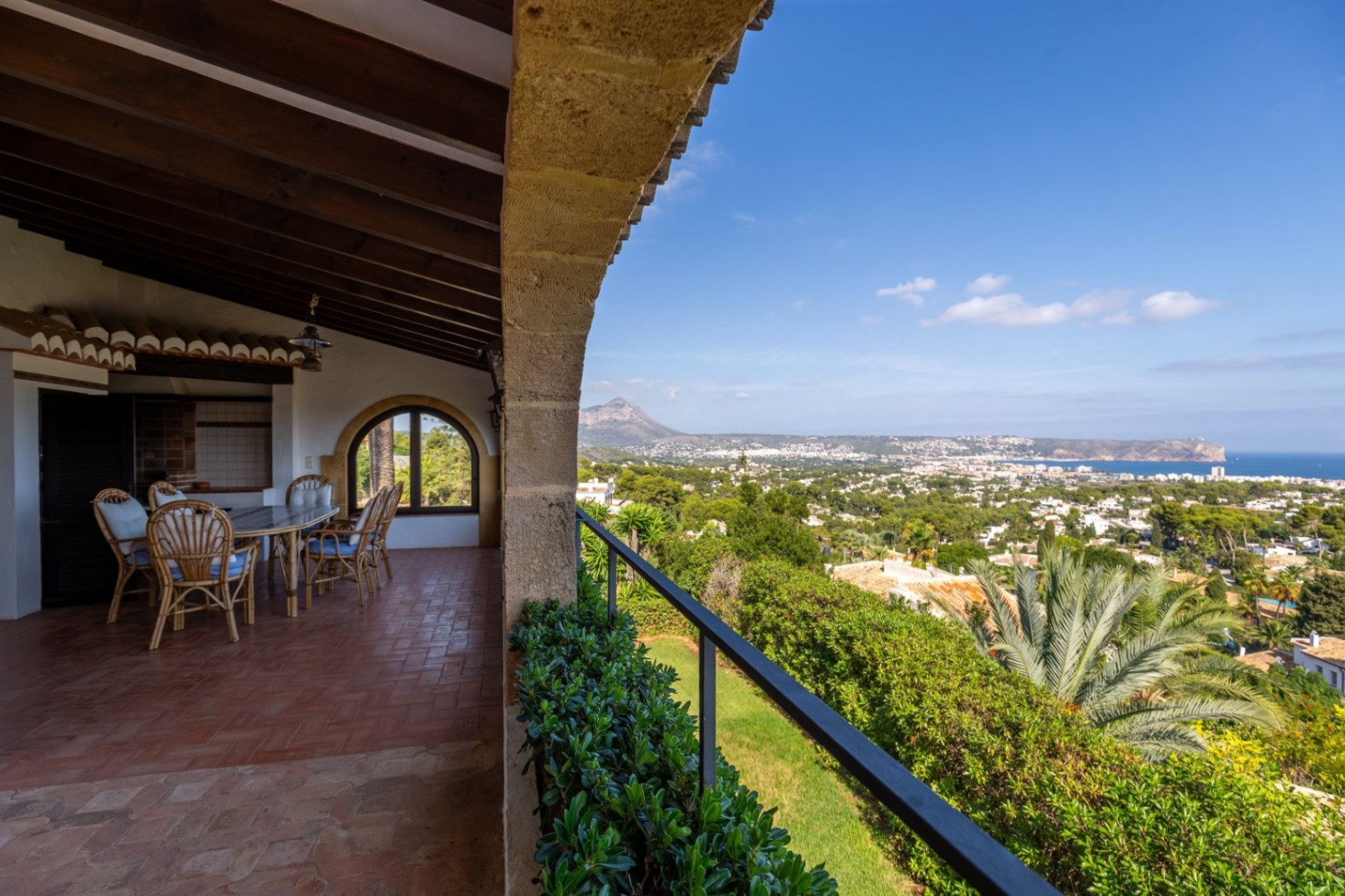 Covered terrace with wood-beamed ceilings and terracotta flooring, featuring a dining area and panoramic views of the coastal valley and sea.