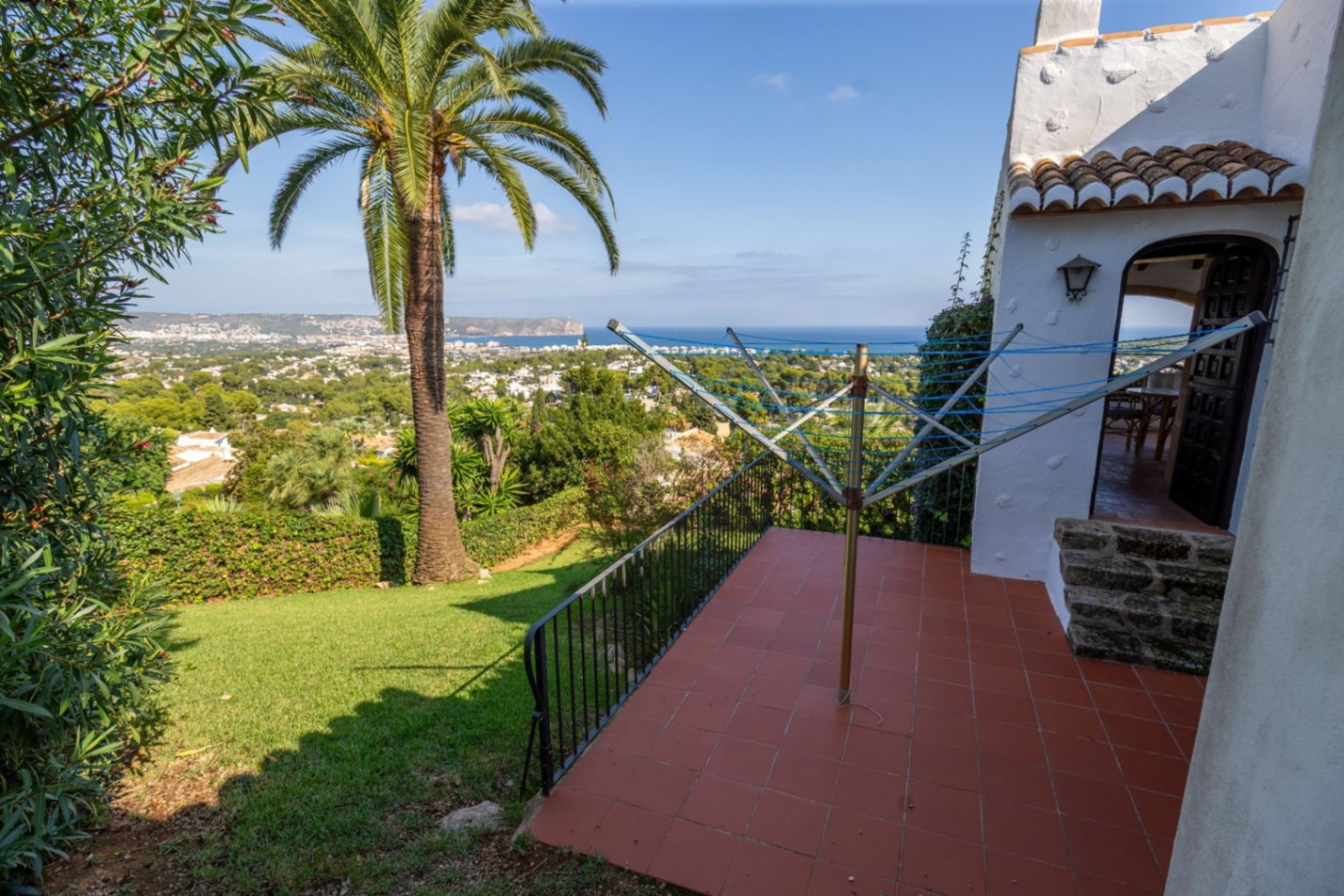 Elevated terrace with terracotta tiles and black railing, offering panoramic views of the coastal valley, the sea, and a mature palm tree.