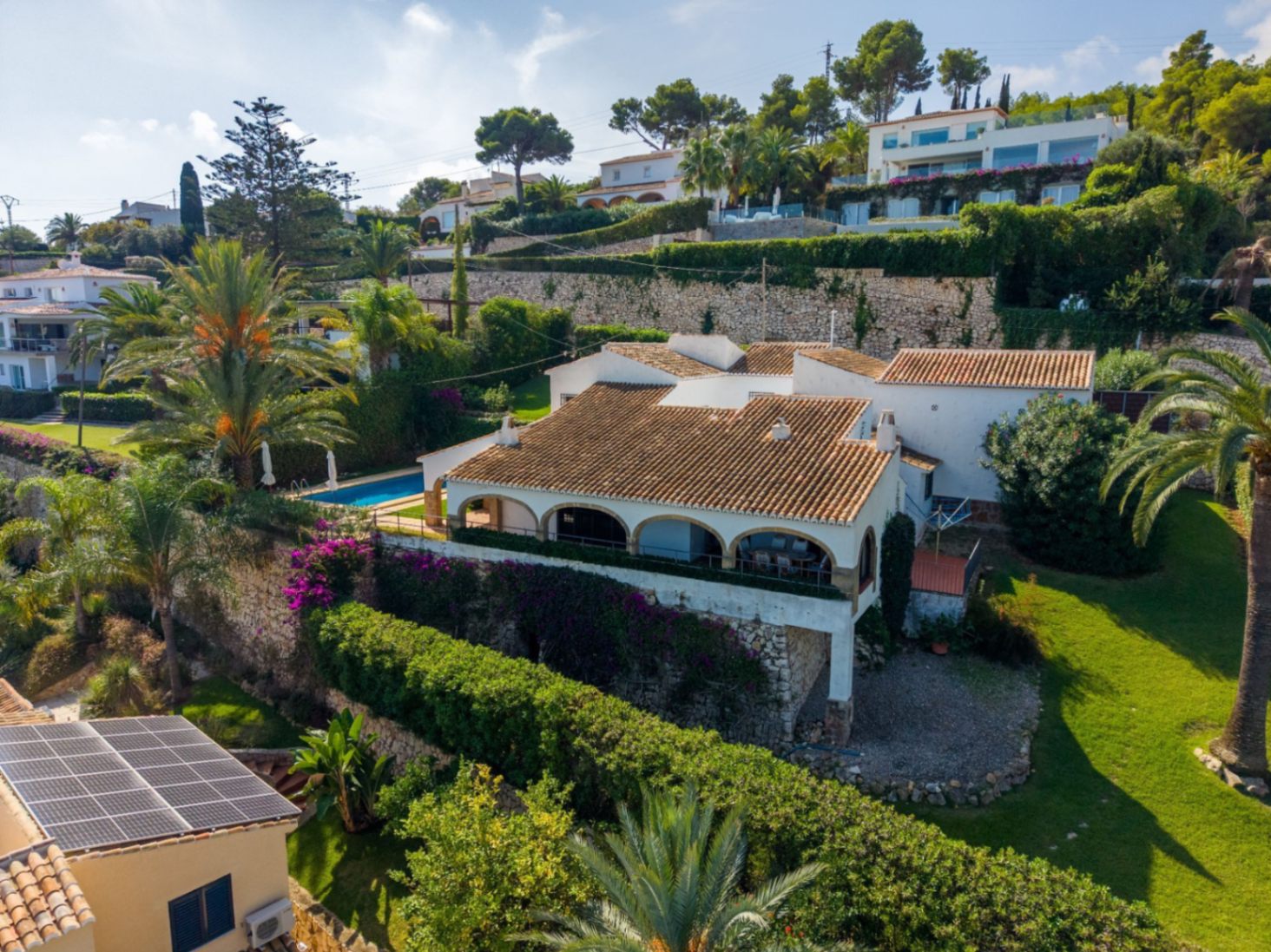 Elevated view of a Mediterranean villa with terracotta roofing, arched terrace, and private pool, set amidst tiered stone walls and lush gardens with palm trees.