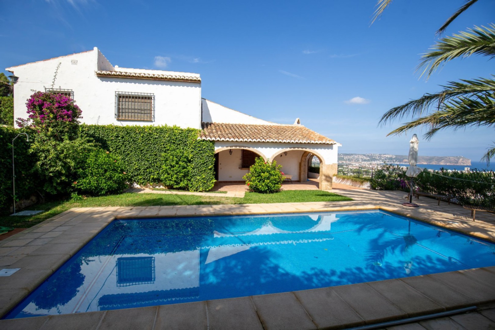 Exterior view featuring a rectangular swimming pool, stone-arched porch, and white facade with climbing bougainvillea overlooking the Mediterranean coast.
