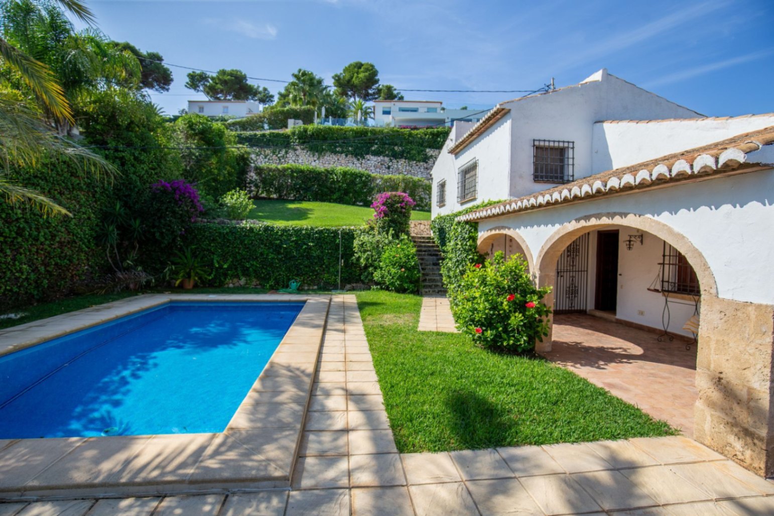 Exterior view of a Mediterranean villa featuring a rectangular pool, arched stone porch, and tiered gardens with mature greenery and bougainvillea.