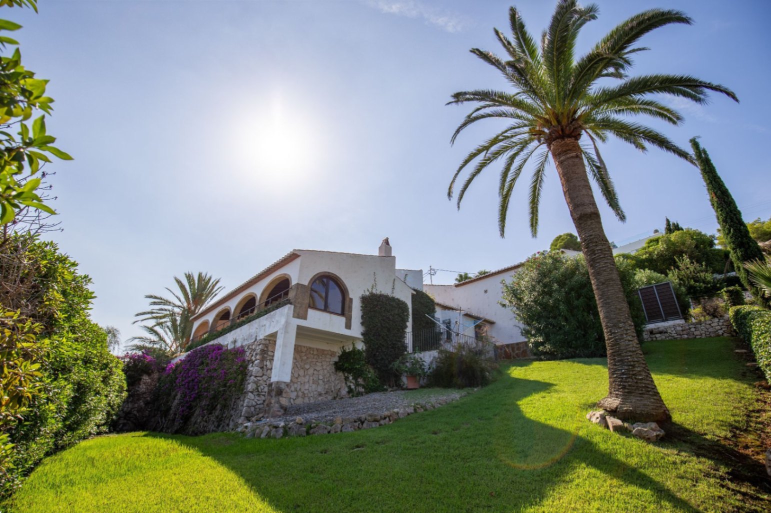 Exterior view of a white Mediterranean villa featuring a traditional arched naya, stone masonry walls, and a sloped lawn with a palm tree.