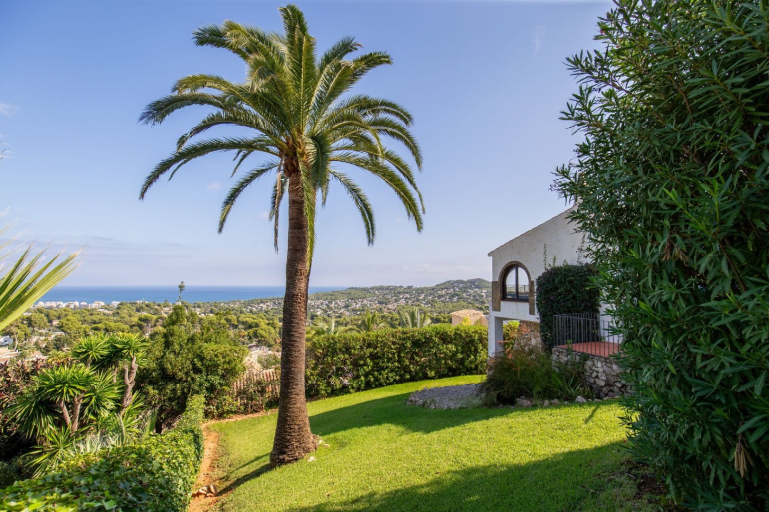 Garden featuring a mature palm tree and lawn, with a view of the white villa's stone facade and arched window. Panoramic coastal and sea vistas in the background.